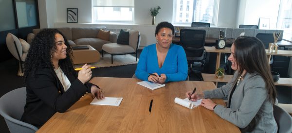 Three women take notes at a conference table in a New York City office.