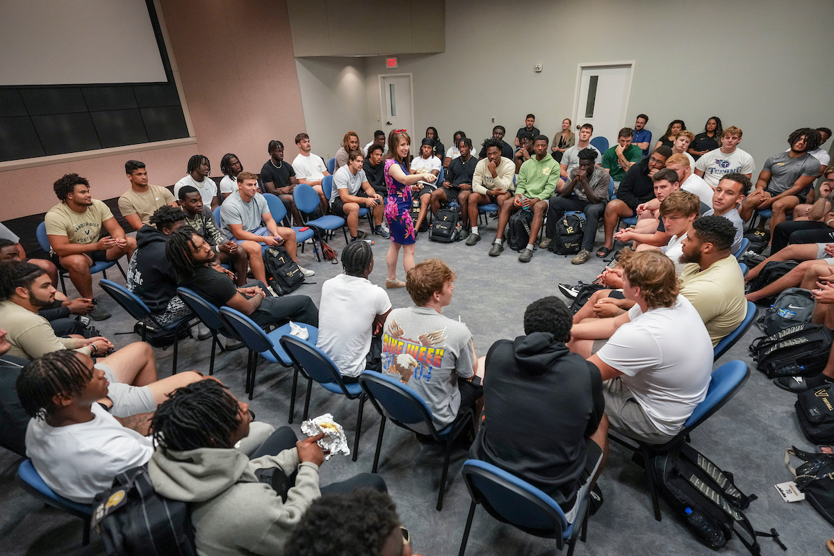 A woman standing in the middle of a circle speaking to a group of men