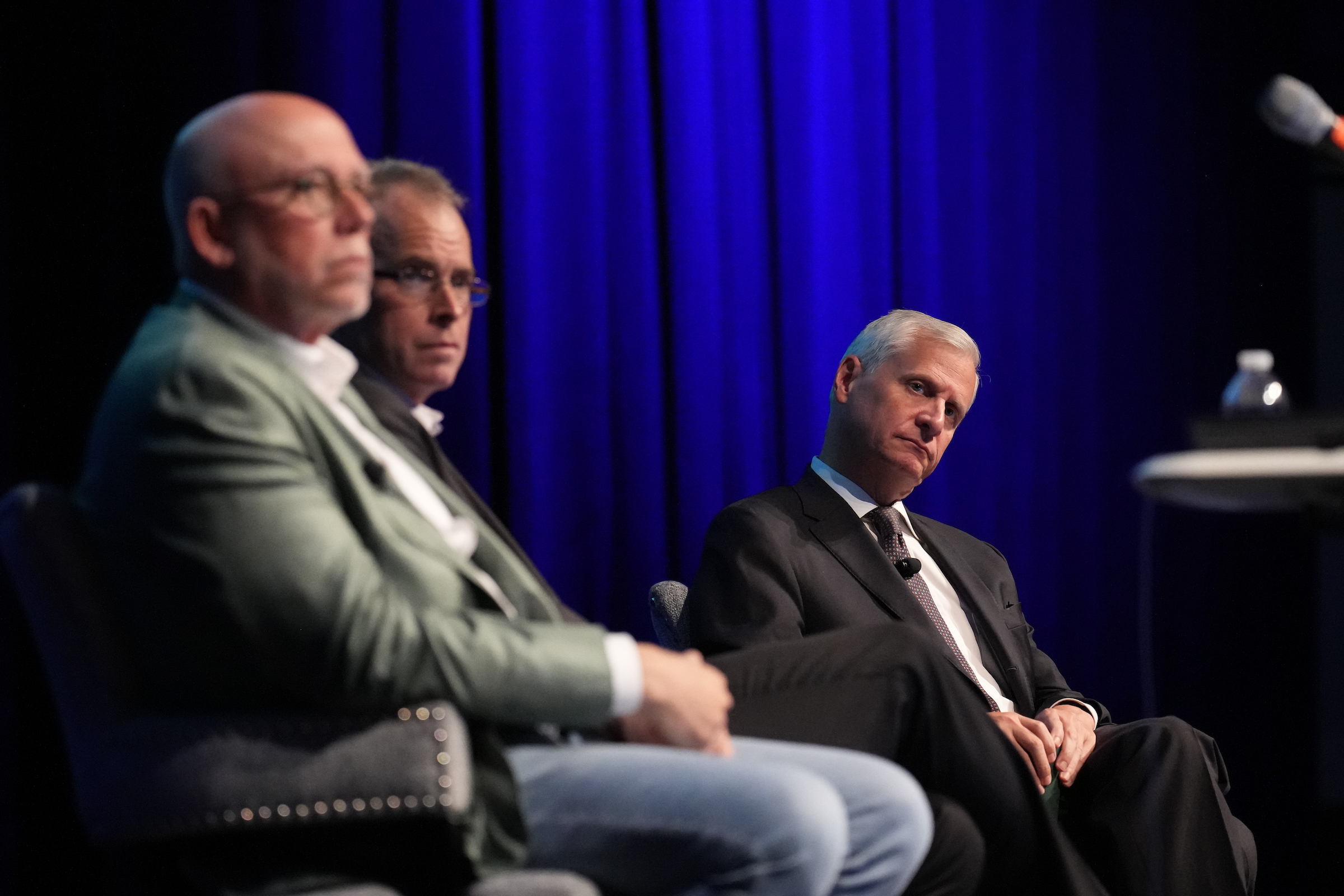 Three men seated next to each other up on a stage with a blue curtain in the background.