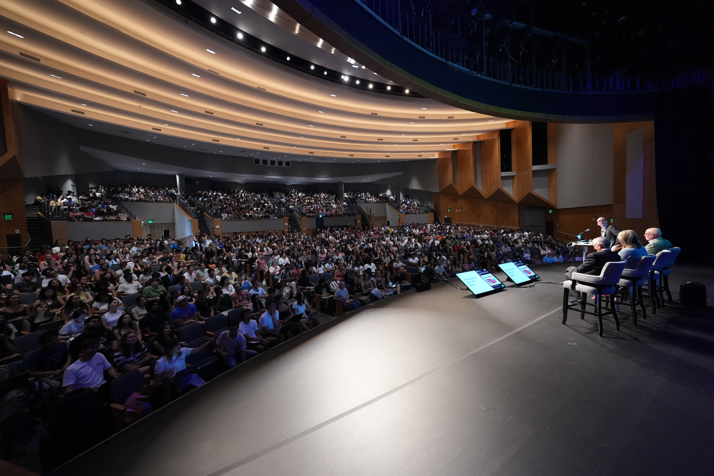 Students sit in an auditorium with seated professors on a stage.