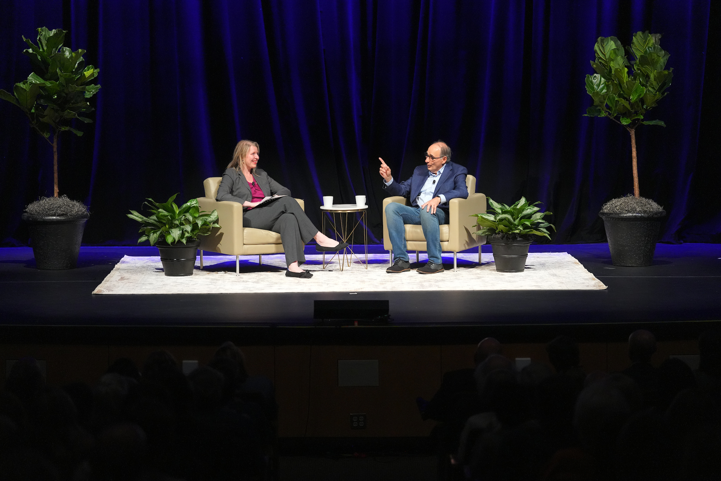 David Axelrod sits with Nicole Hemmer on a stage with a blue curtain in the background