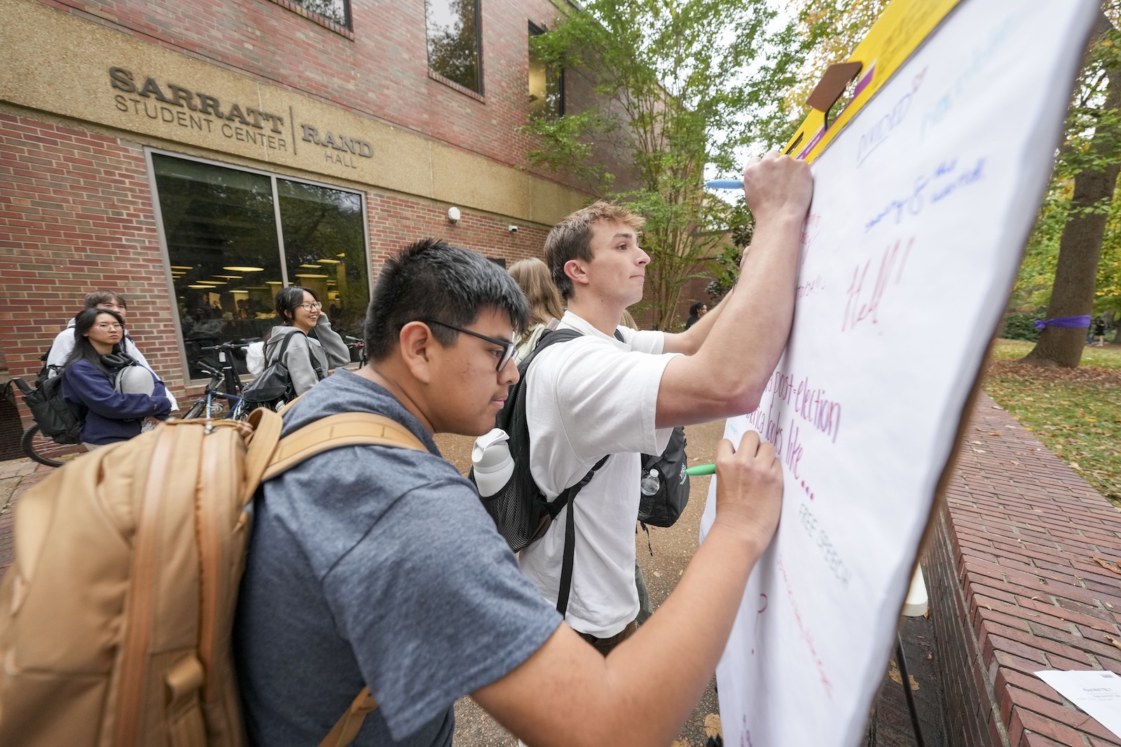 Two males write a message on a large pad of paper outside of a dining hall.