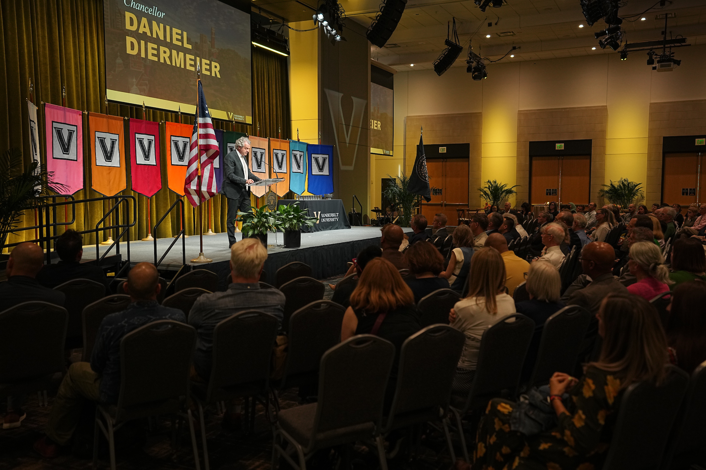 A crowd sits before a podium with various colored Vanderbilt banners and a speaker in front of them. 