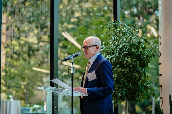 Dean McNamara speaking at a podium in front of large glass walls with the view of trees outside. 