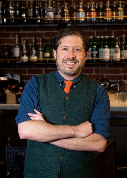 Portrait of a man in a vest in front of a bar with many bottles of spirits behind him.