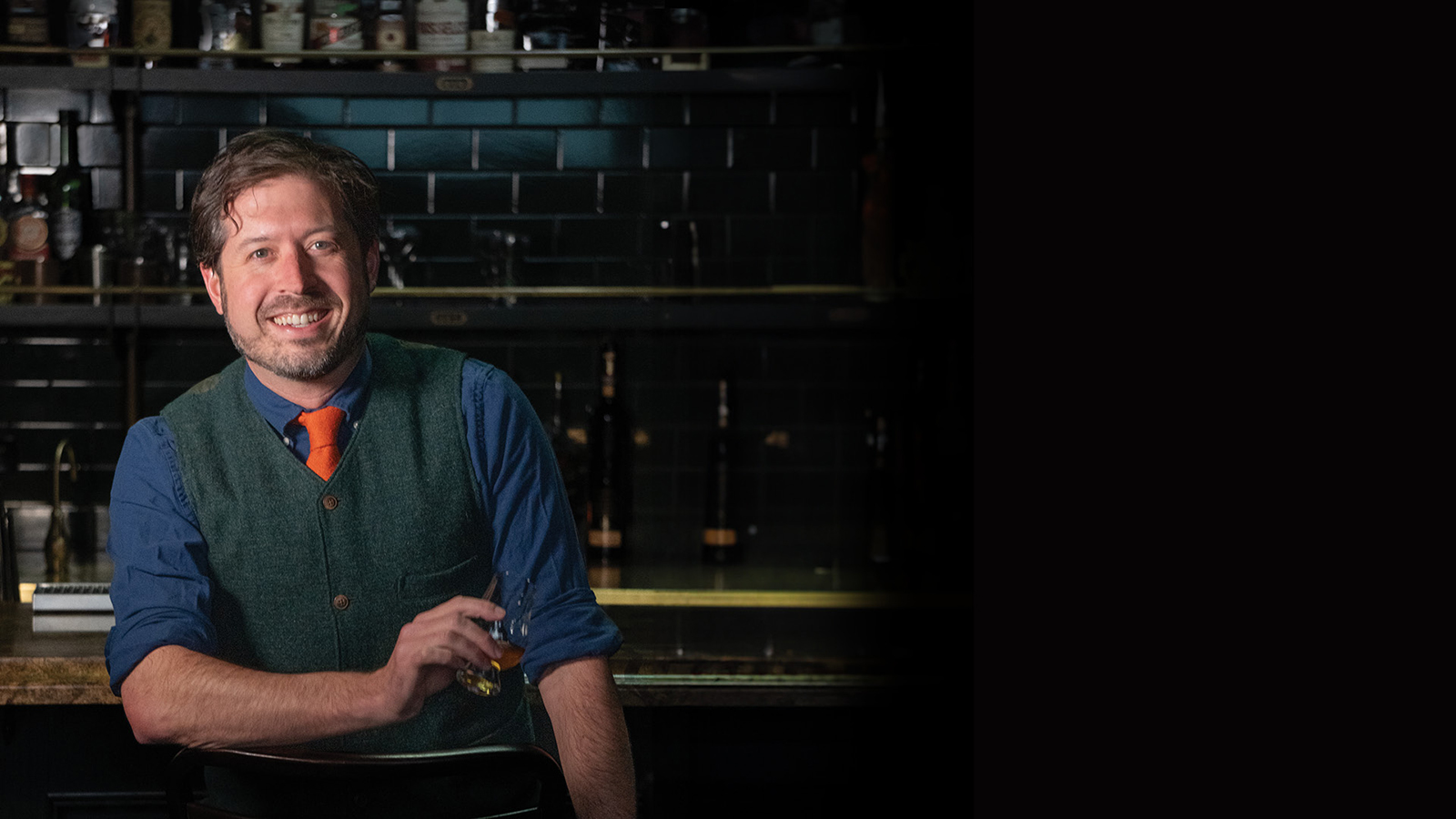 Vanderbilt alumnus Jacob Grier standing at the bar of a Portland, Oregon, bar with many bottles of spirits behind him.