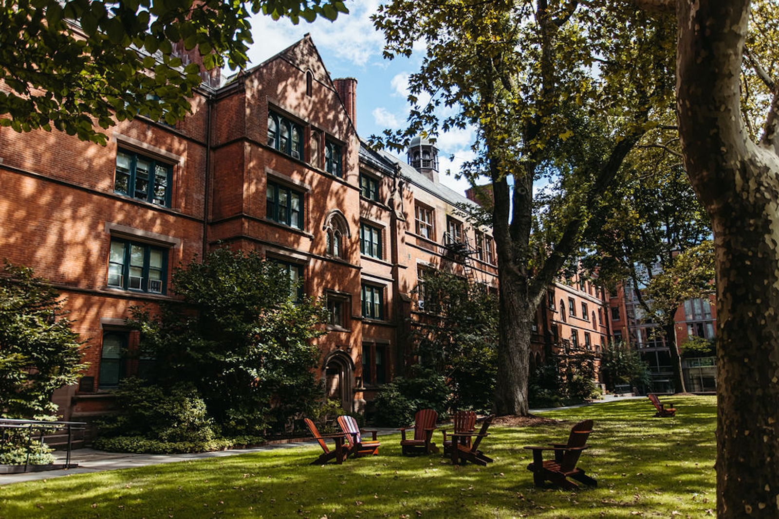 An exterior shot of a brick building surrounded by mature trees and a lawn with several Adirondack chair. (Courtesy of The General Theological Seminary)