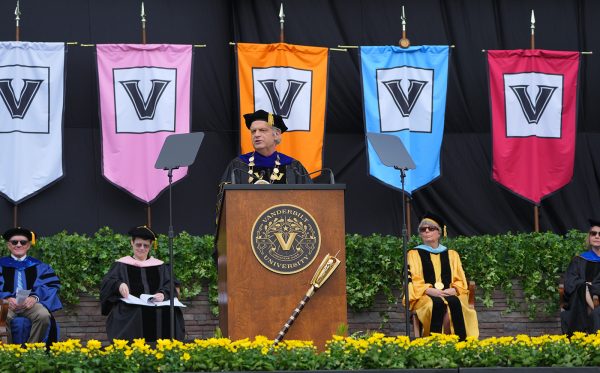 Chancellor Daniel Diermeier speaking from the podium on the main Commencement stage at GEODIS Park with color school banners behind him.