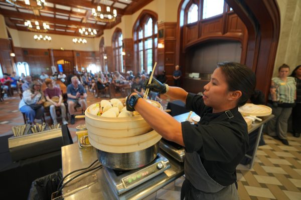 A woman shifts buns in a stacked bamboo steamer in front of tables filled with seated people.