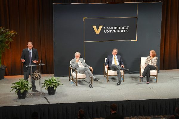 A speaker stands at a podium with three seated participants by his side. In the background is a Vanderbilt University banner.