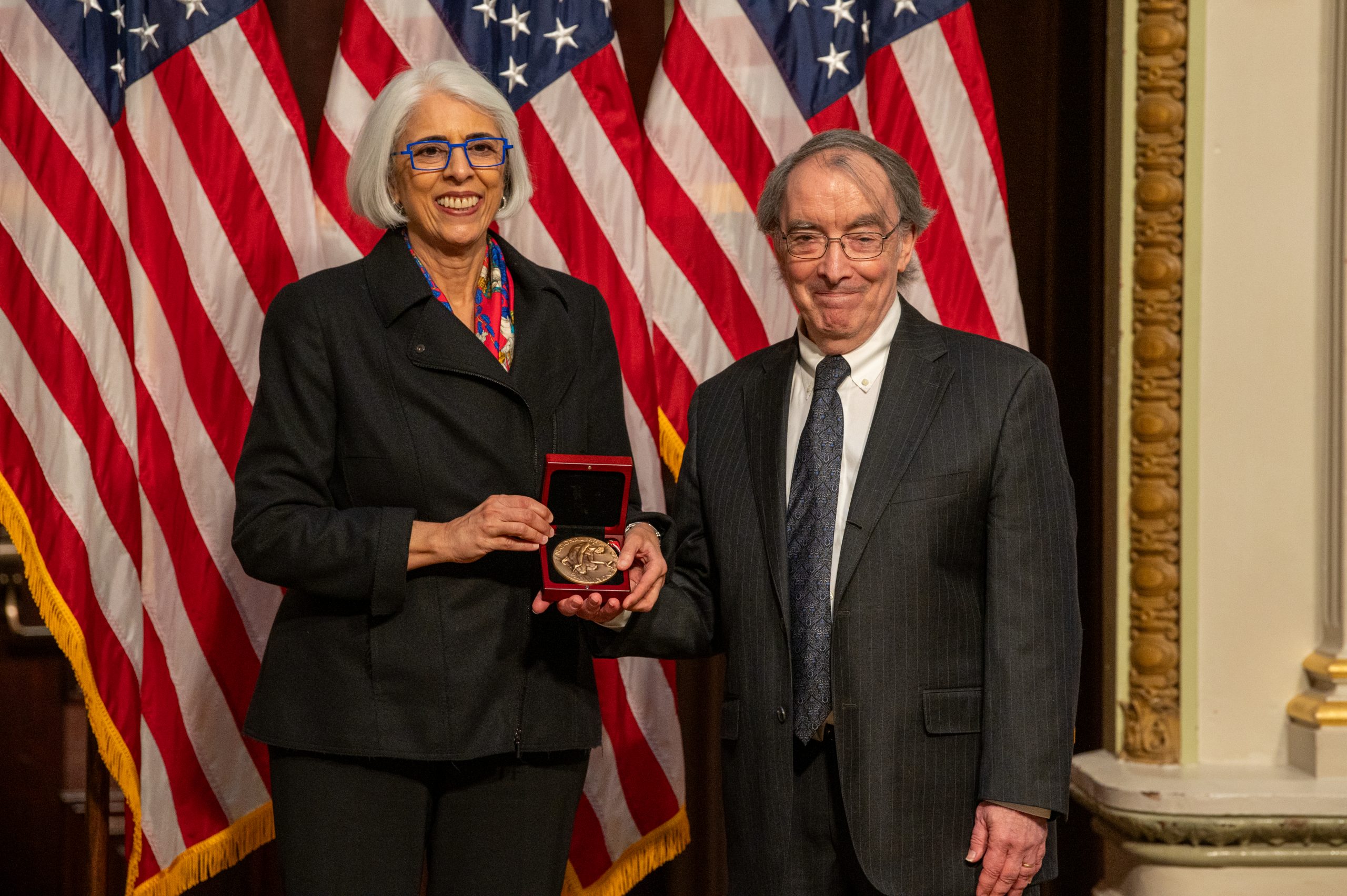 Larry Bartels holds a medal with Arati Prabhakar and three U.S. flags as the backdrop 