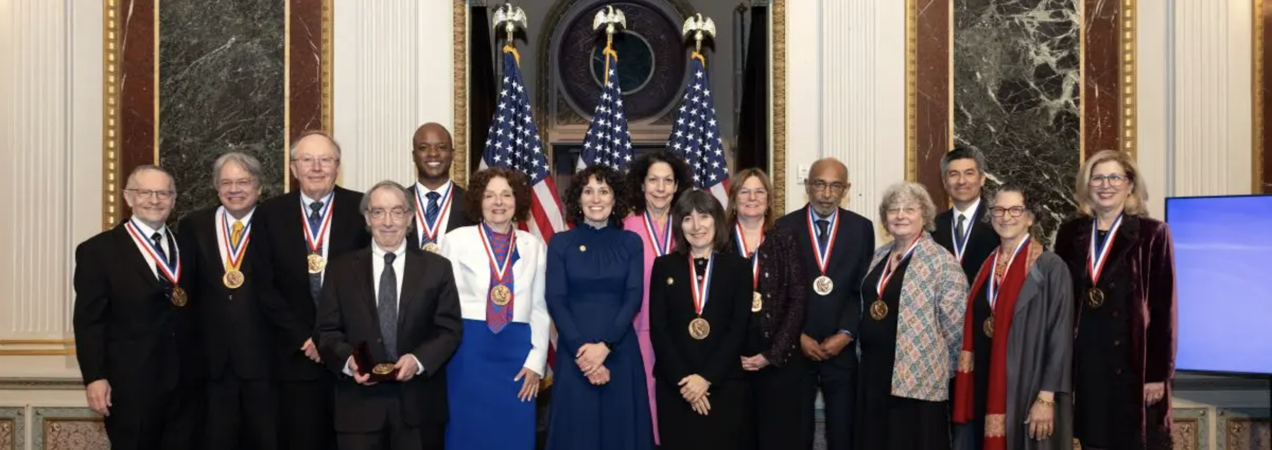 National Medal of Science winners stand together in a group in front of three U.S. flags and marble columns