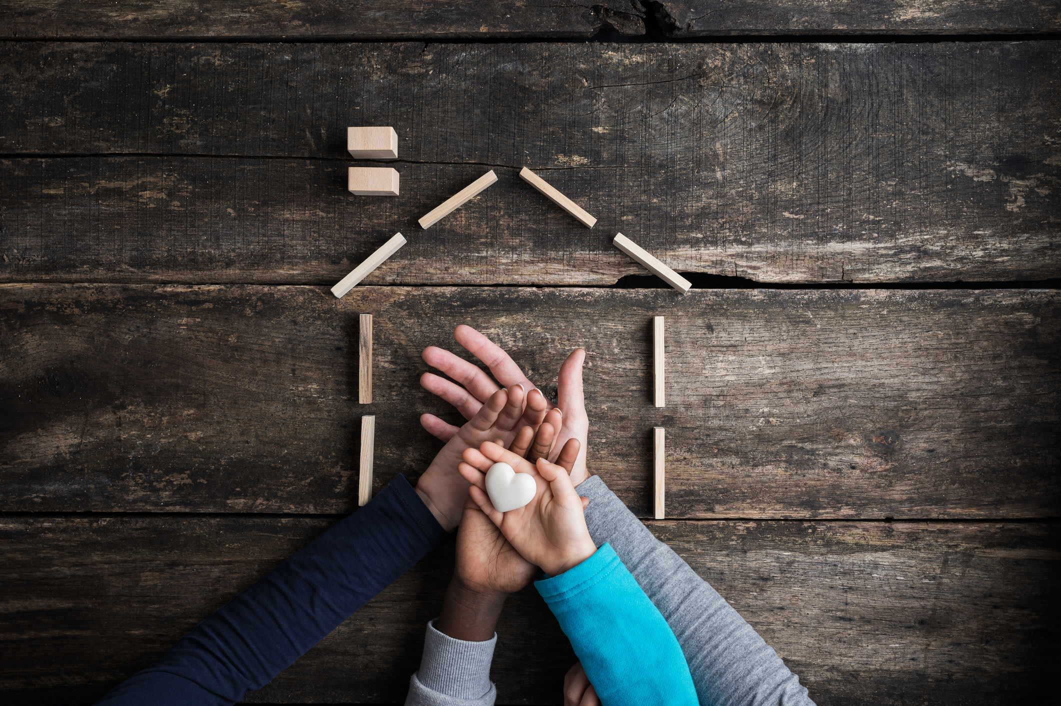 multiple hands holding white heart inside model of house framed with wooden sticks