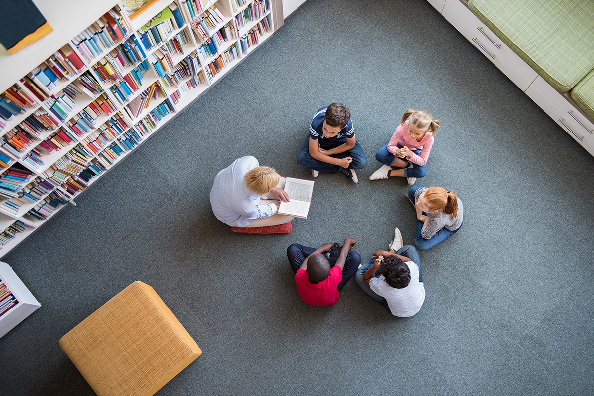 Top view of librarian sitting with five multiethnic children on floor. Teacher reading book to girls and young boys at school.